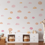 A child's room with a white wall adorned with pink and orange pastel cloud decals, a stuffed blue whale on a wicker basket, and a tall giraffe toy next to a white storage unit.