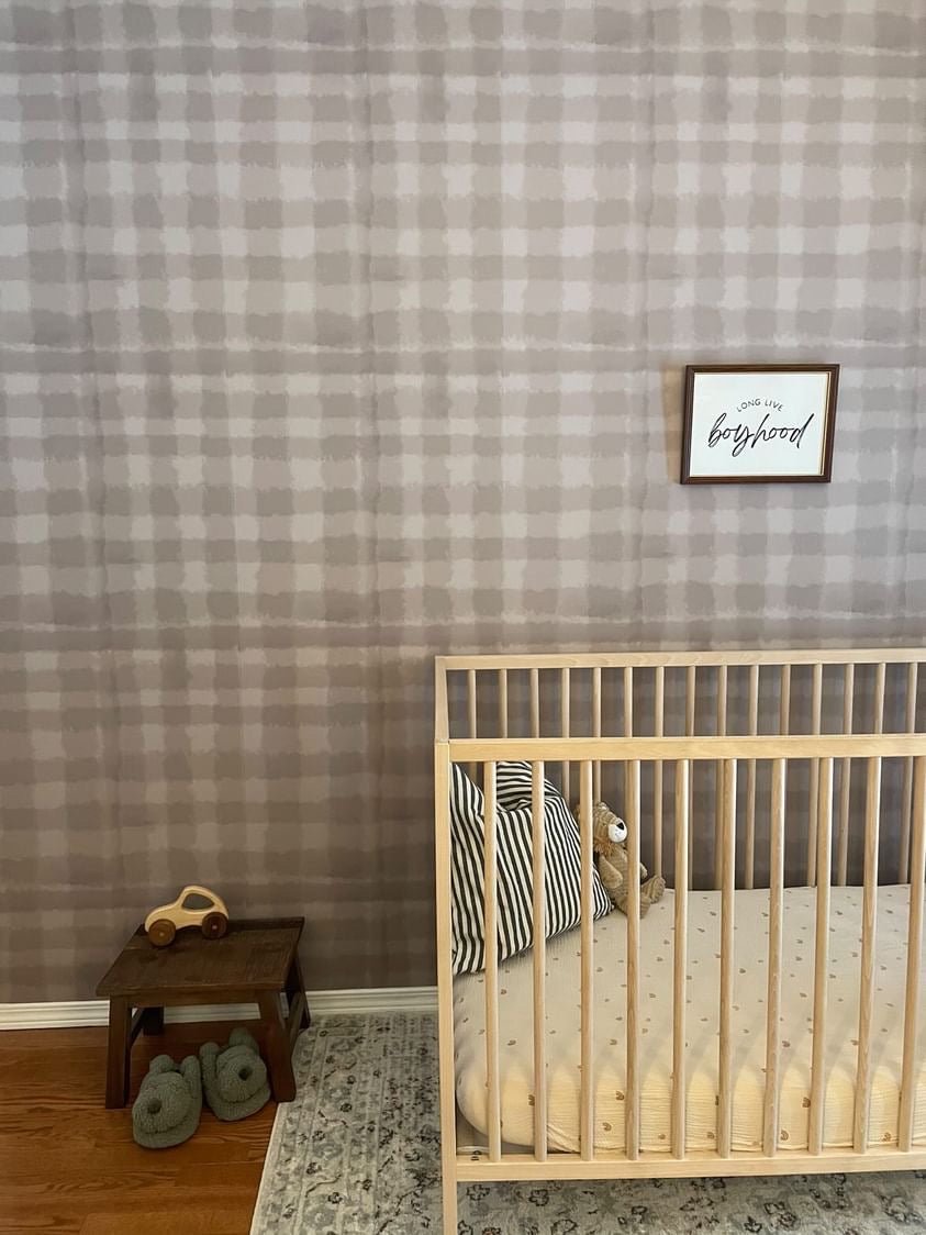A cozy nursery with a wooden crib and plush toy, wooden stool with a car toy, knitted booties, and a "long live boyhood" wall frame.