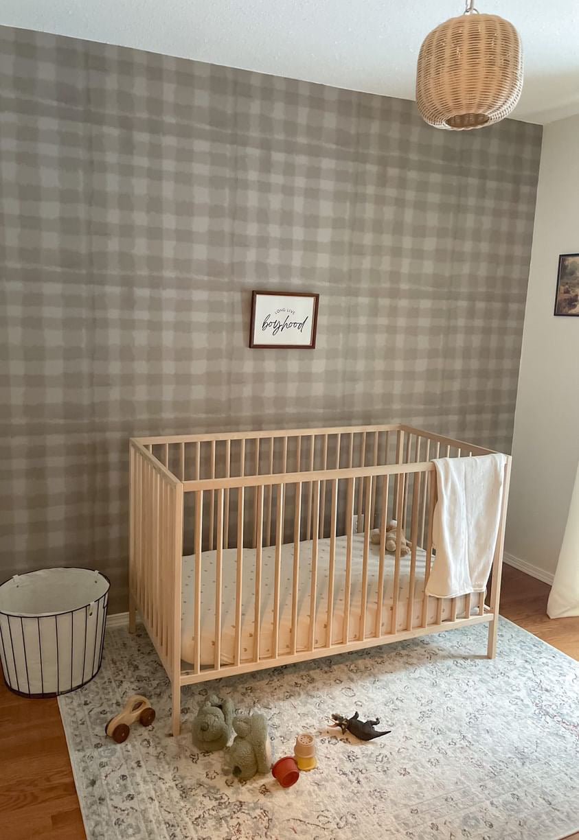 Airy baby room with crib, white blanket, toys, "long live boyhood" frame, rattan light, and gingham wallpaper.