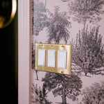 A light switch with three white switches on a brass plate against a toile wallpaper with pink trees and foliage.