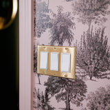 A light switch with three white switches on a brass plate against a toile wallpaper with pink trees and foliage.