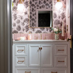 A bathroom vanity with white cabinets, brass handles, and a pink toile tree-pattern wallpaper.