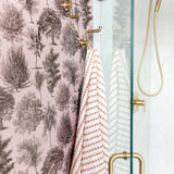 A shower with pink toile tree-pattern wallpaper, brass hooks holding striped towels, and a glass door.