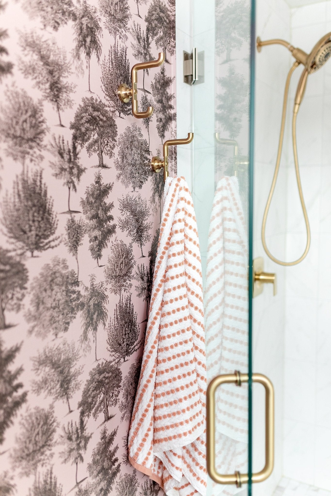 A shower with pink toile tree-pattern wallpaper, brass hooks holding striped towels, and a glass door.