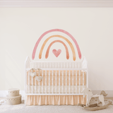 A nursery with a large pastel rainbow wall sticker positioned above a classic white crib. The crib has a beige blanket, and nearby is a white rocking horse toy. The floor is adorned with a soft rug and there's a pair of decorative white hearts hanging on the crib.