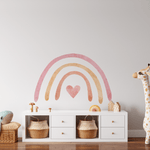 A children's playroom with a pastel rainbow wall sticker above a white dresser. The room has a whimsical feel with a large plush dolphin toy on a wicker bench, a giraffe-shaped toy, and woven baskets for storage.