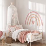 A cozy corner of a child's bedroom featuring a pastel rainbow wall decal. The room includes a white canopy over a single bed with pink bedding, a fluffy white rug on the floor, and a small wooden desk with a red chair.