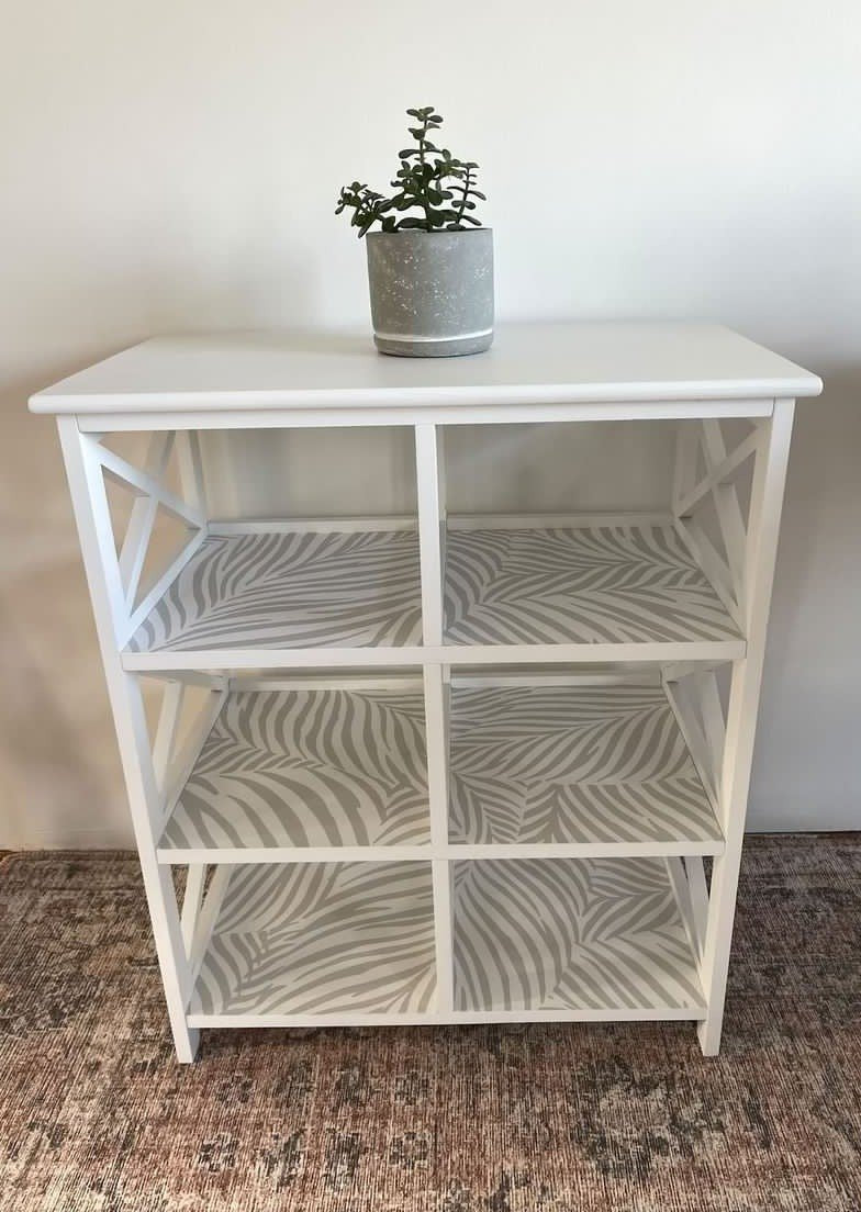 This photo shows a white open faced shelf with grey and white Scandinavian style wallpaper lining each shelf.