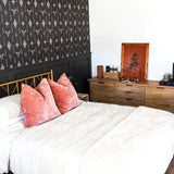 A wide-angle view of the bedroom focused on the cohesive design featuring the Western wallpaper and dark wainscoting. A gold metal bed frame sits against the feature wall, adorned with white bedding and coral accent pillows. A wooden dresser opposite the bed holds a framed picture, decorative items, and a stack of books, while the chandelier from the first image is visible, maintaining the room's contemporary Western aesthetic