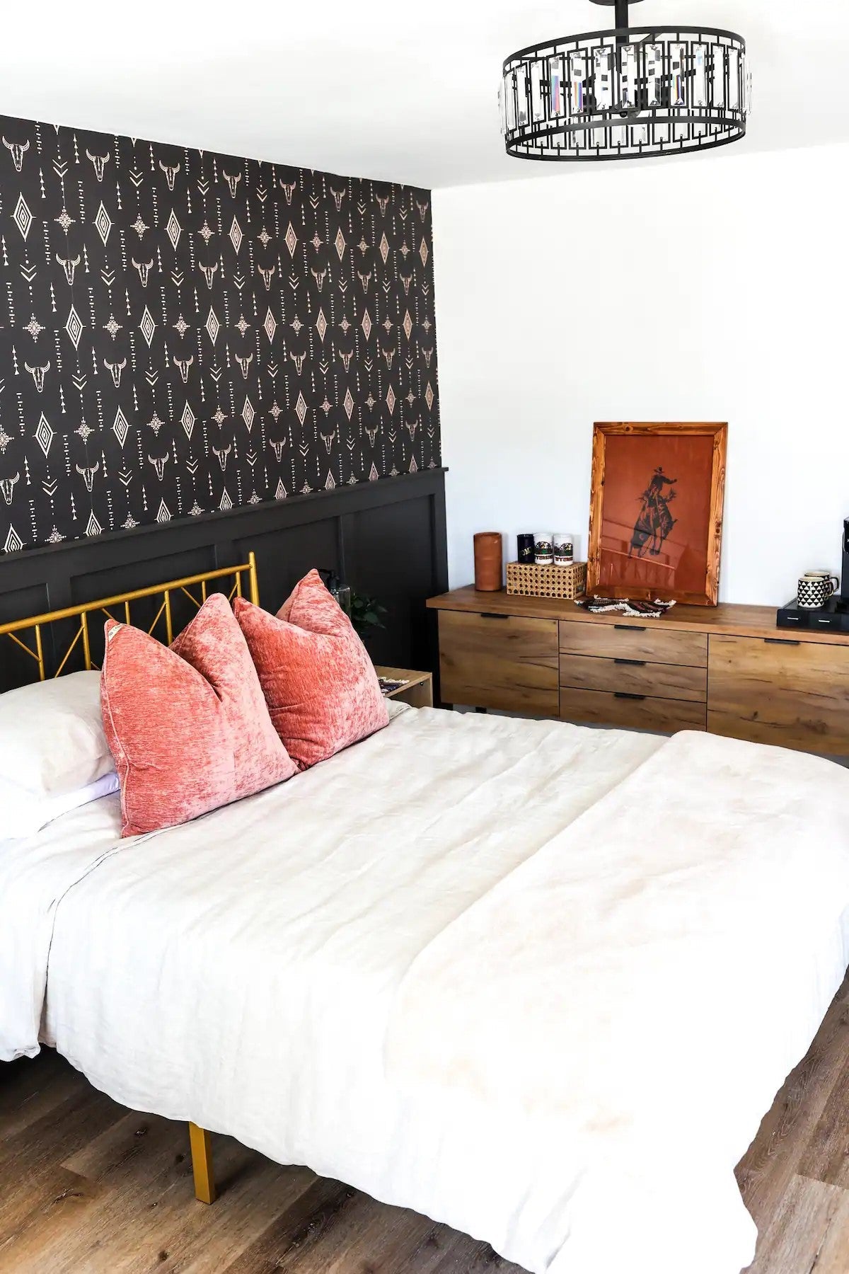 A wide-angle view of the bedroom focused on the cohesive design featuring the Western wallpaper and dark wainscoting. A gold metal bed frame sits against the feature wall, adorned with white bedding and coral accent pillows. A wooden dresser opposite the bed holds a framed picture, decorative items, and a stack of books, while the chandelier from the first image is visible, maintaining the room's contemporary Western aesthetic