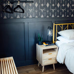Another angle of the bedroom highlighting the Western-inspired wallpaper complemented by a dark blue paneled wall below. Beside the bed with crisp white linens is a natural wood bedside table with rattan drawer fronts, topped with a modern lantern-style lamp, books, and a small potted plant.