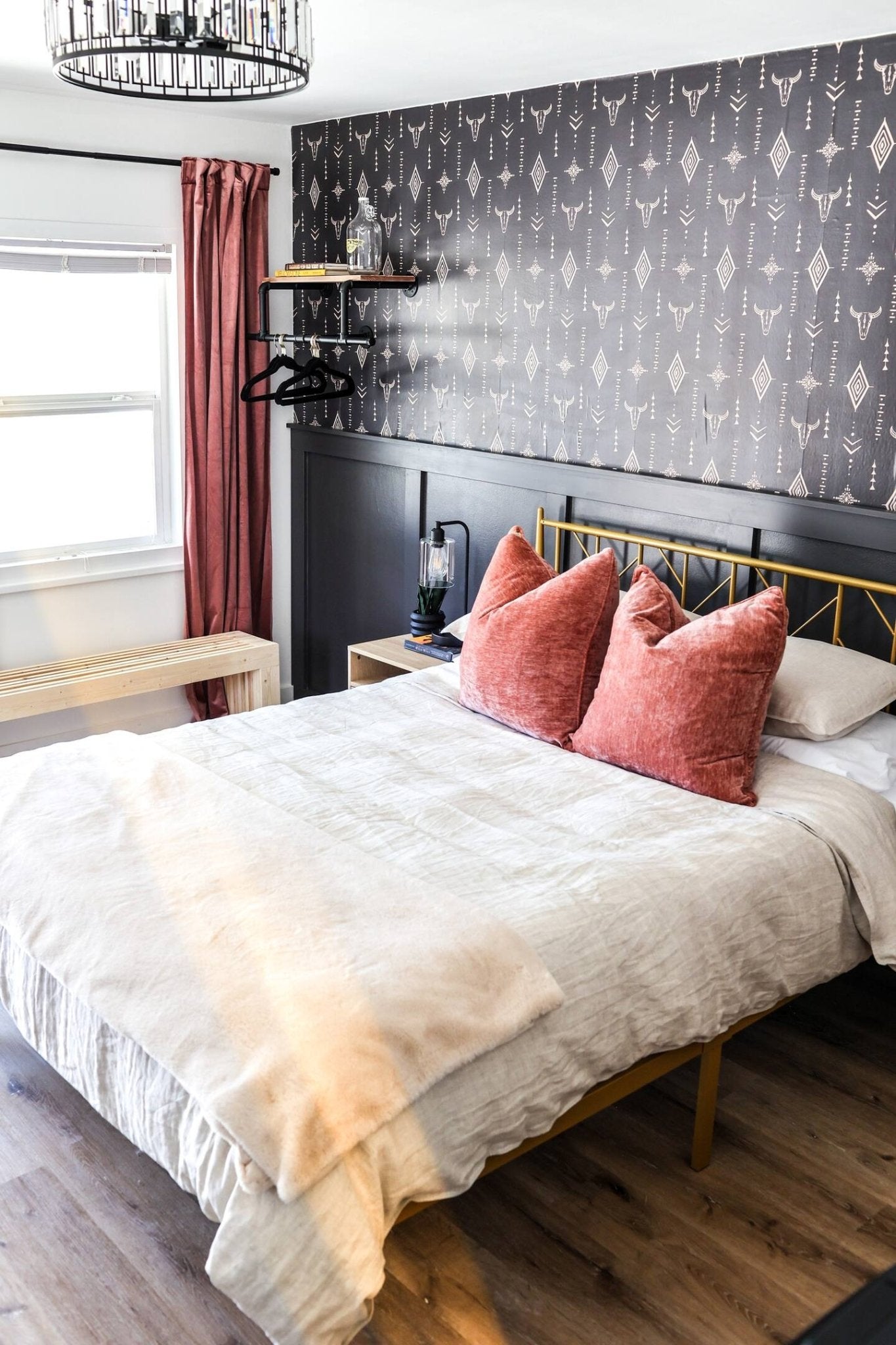 A stylish bedroom showcasing a bold Western-themed wallpaper with white illustrations of arrows, diamonds, and bull skulls on a dark background. The room features a gold metal bed frame, white bedding with coral throw pillows, and a beige throw at the foot of the bed. A dark paneled wall anchors a floating wooden shelf and a black modern chandelier adds a touch of elegance