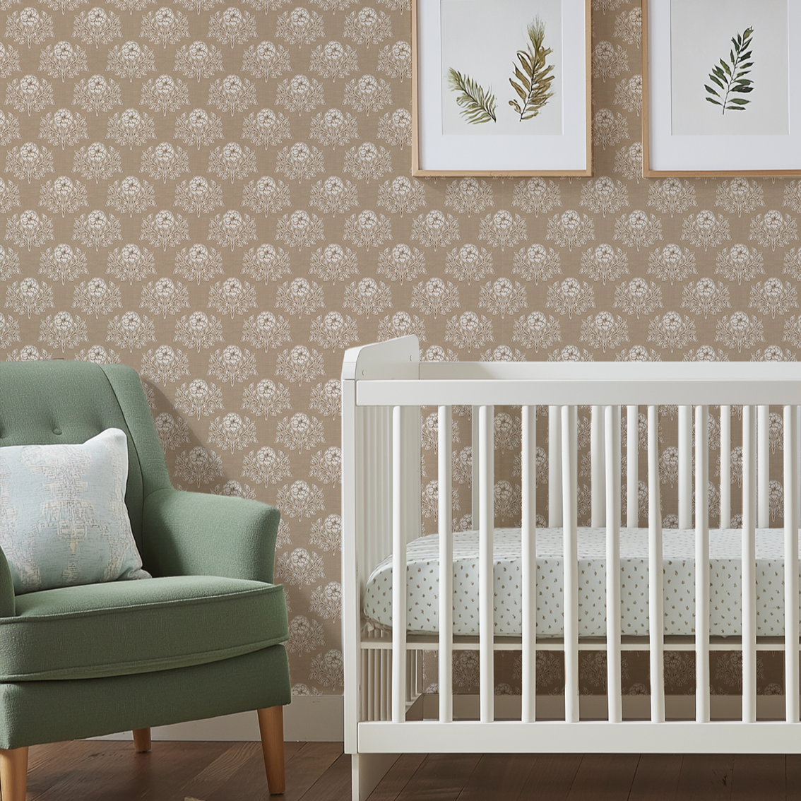 A styled nursery corner featuring a white wooden crib with a patterned mattress, a green upholstered armchair with a white pillow, and a wall covered in beige damask-patterned wallpaper with two framed botanical prints above.