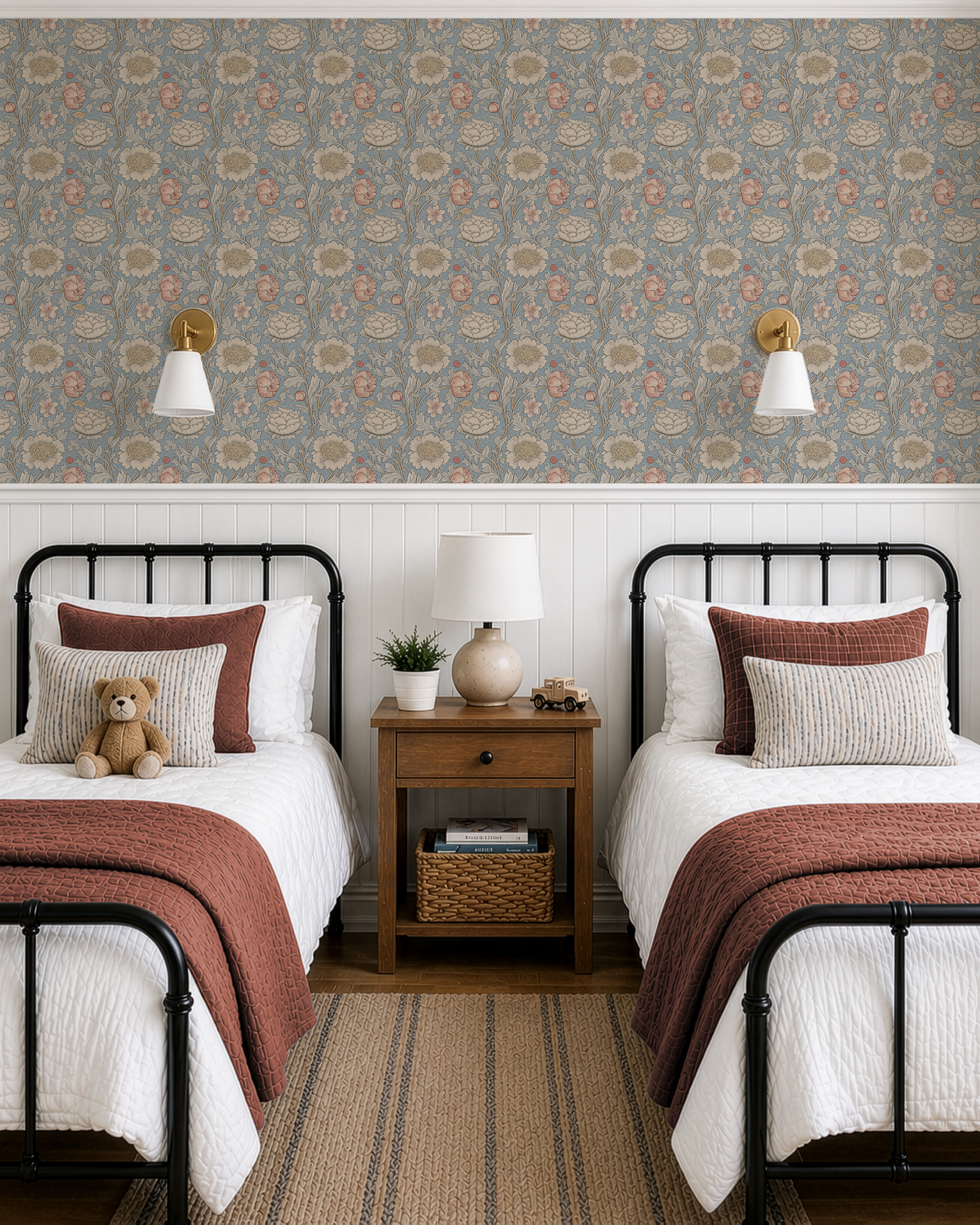 A styled children's bedroom featuring a prominent blue floral wallpaper on the upper wall, white wainscoting, two single beds with white bedding and rust-red throws, a wooden nightstand, and a teddy bear.