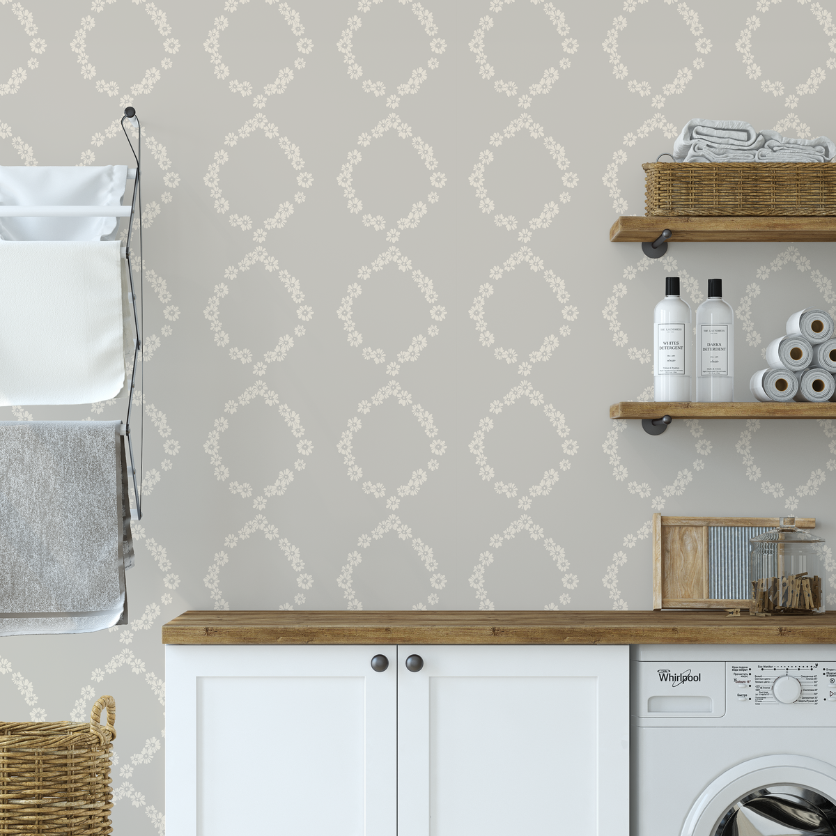 A styled laundry room corner featuring light beige wallpaper with a repeating white floral trellis pattern, a wooden countertop, white cabinets, a washing machine, and wall-mounted shelves with towels and laundry bottles.