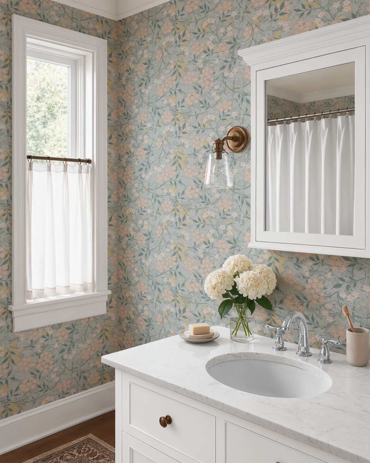 A bathroom interior featuring floral wallpaper in muted sage green, beige, and soft pink tones, a white vanity with a marble countertop, a white sink, a brass wall sconce, a window with sheer curtains, and a mirror.
