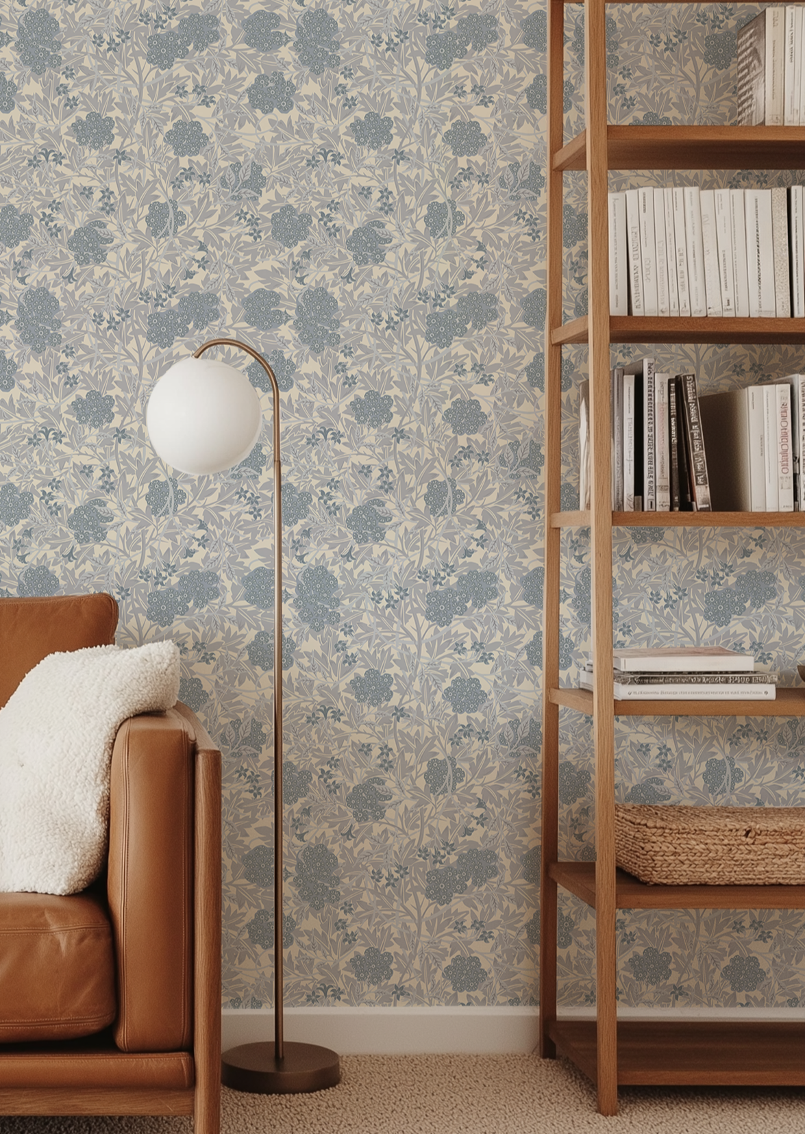 A living room corner featuring a blue and beige floral patterned wallpaper, a brown leather armchair with a white throw pillow, a brass floor lamp with a white globe shade, and a tall wooden bookshelf.