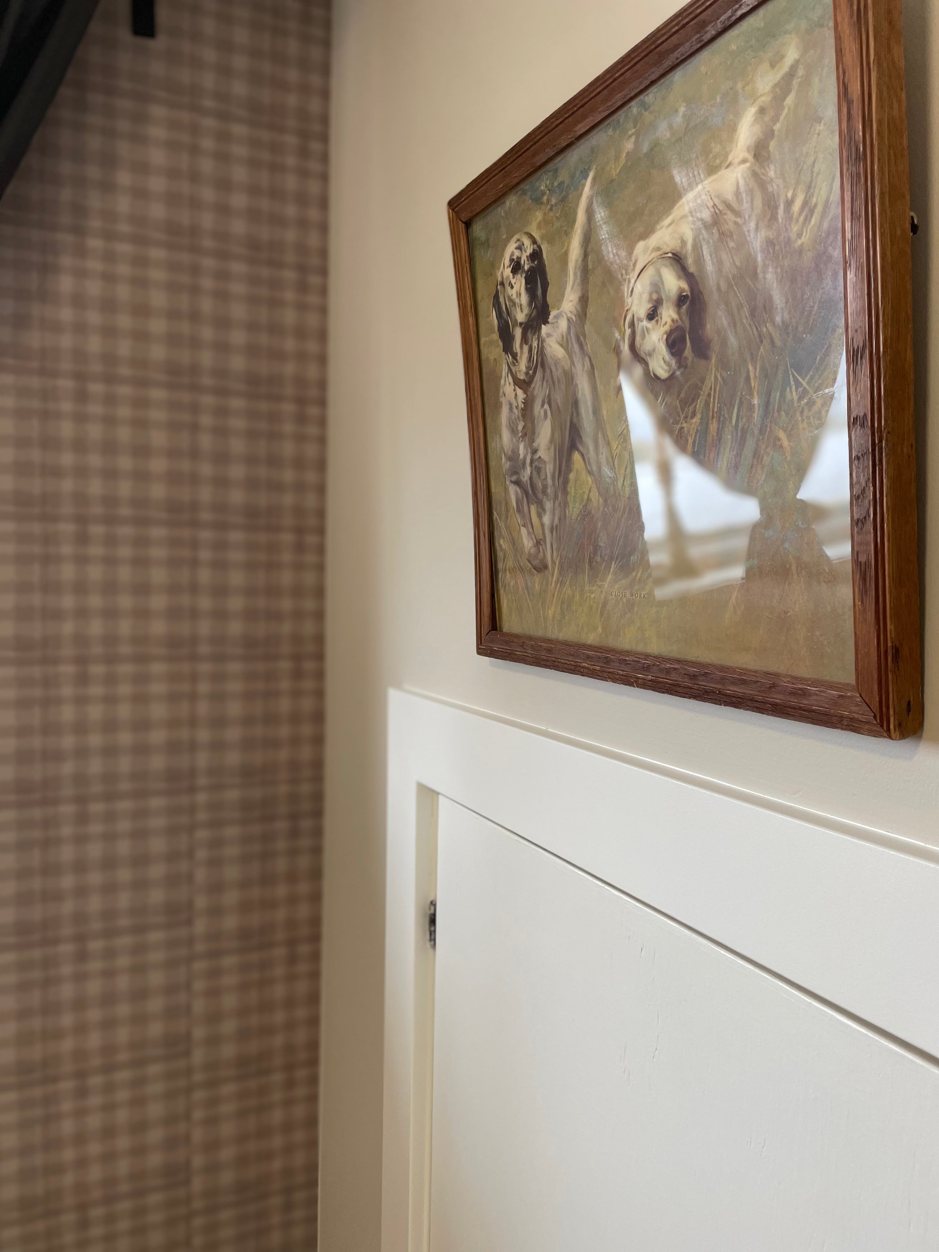 A painting of two dogs running in a field, displayed against a mini plaid wallpaper in a cozy room corner