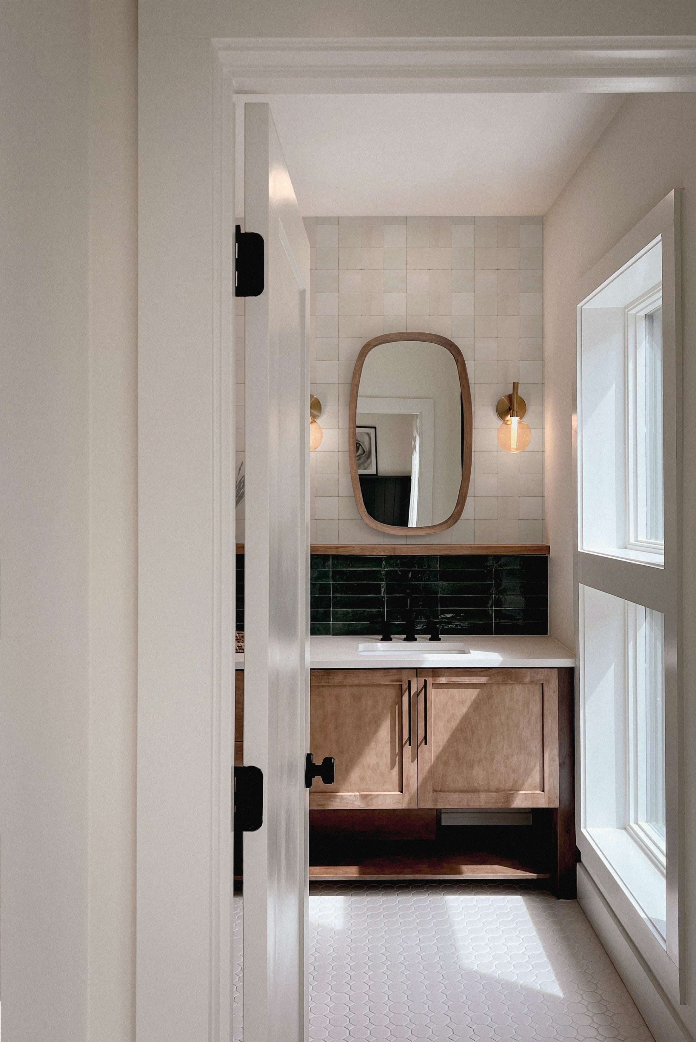View through doorway showing beige zellige tile wallpaper above bathroom vanity with mirror and moody green tile contrast