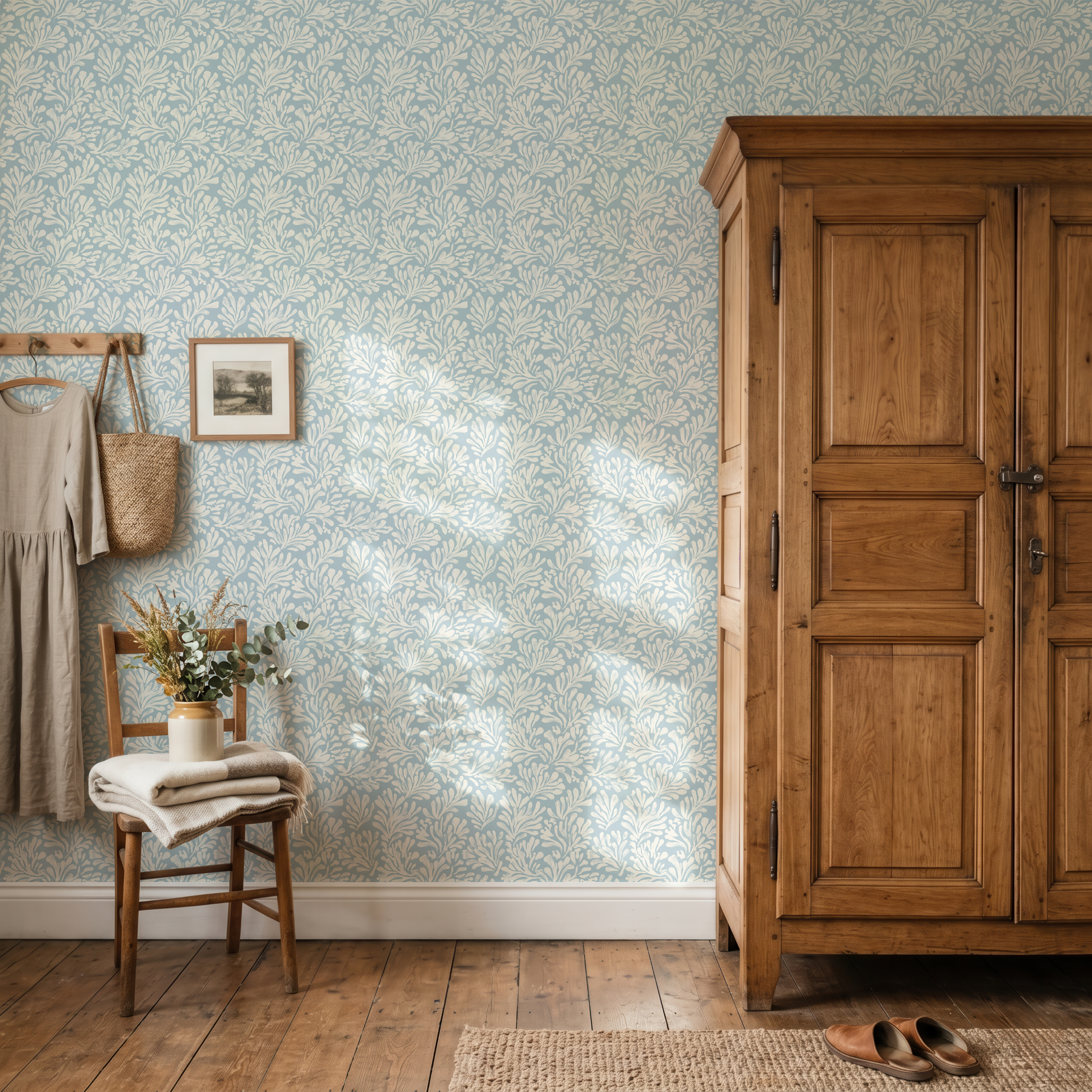 A styled interior room scene showing a light blue and white floral patterned wallpaper on a wall, alongside a large wooden wardrobe, a small wooden chair with folded beige textiles, a vase of dried flowers, and a pair of brown shoes on a woven rug.
