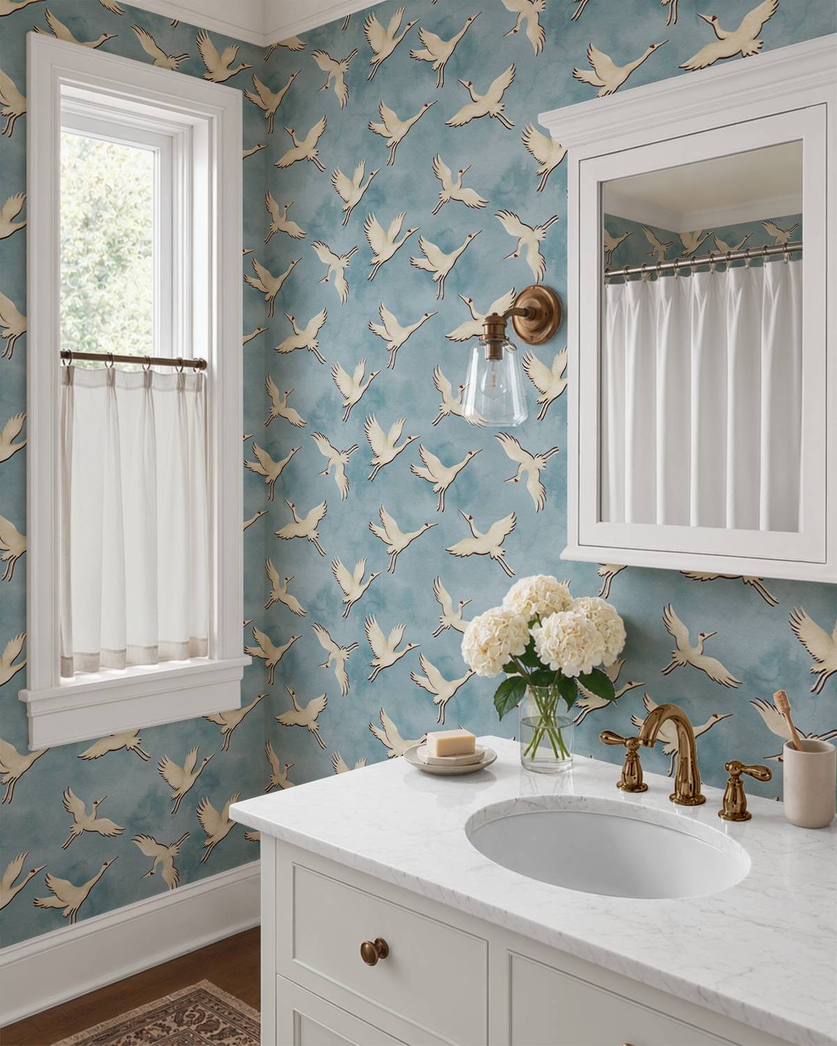 A styled bathroom corner featuring blue wallpaper with a beige crane pattern, a white marble vanity, brass fixtures, a window with sheer curtains, and a wall-mounted mirror.