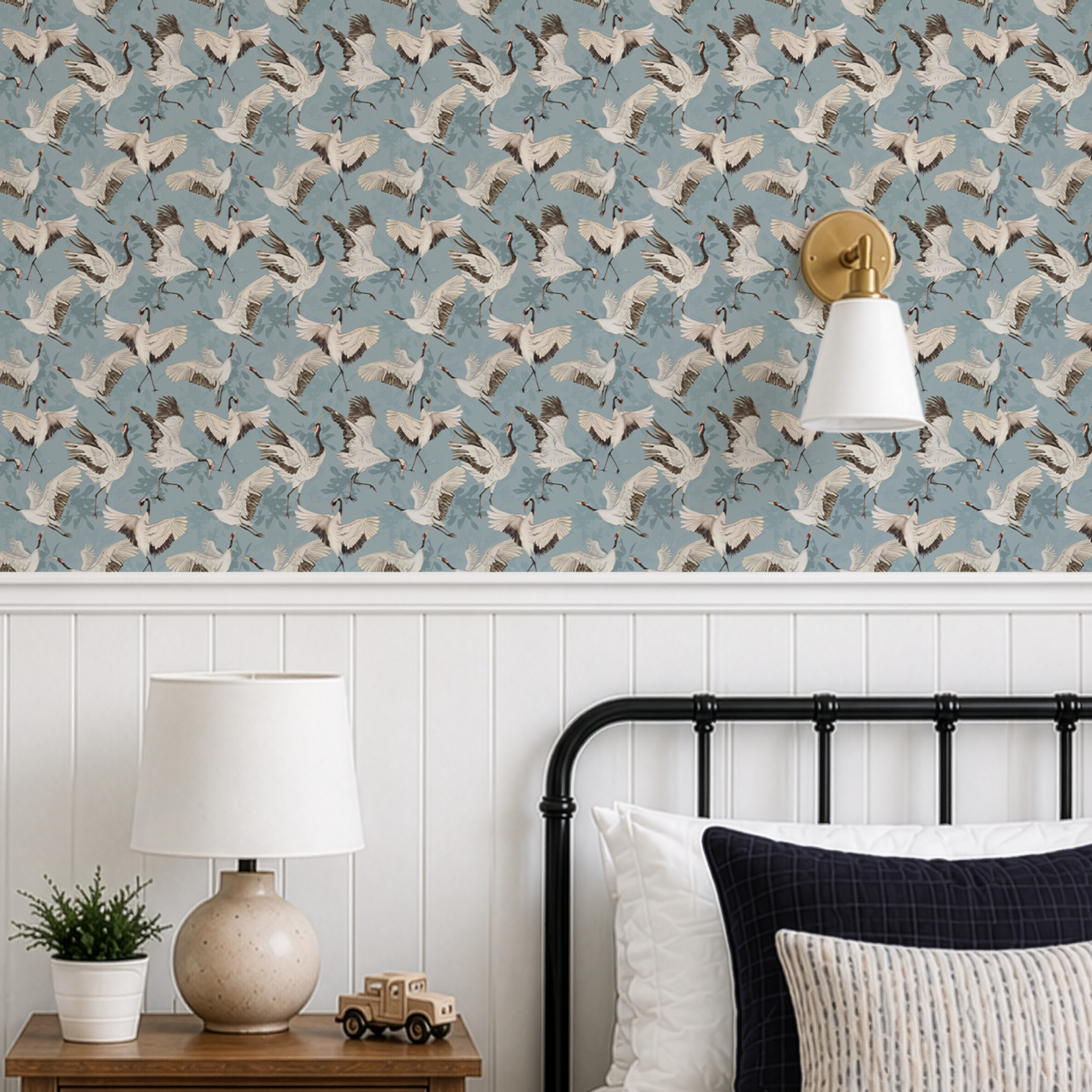A styled bedroom corner featuring wallpaper with a repeating pattern of white and gray cranes on a dusty blue background, installed above white wainscoting. A black metal bed frame with white and navy pillows is visible, along with a wooden nightstand holding a ceramic lamp, a small plant, and a toy truck.