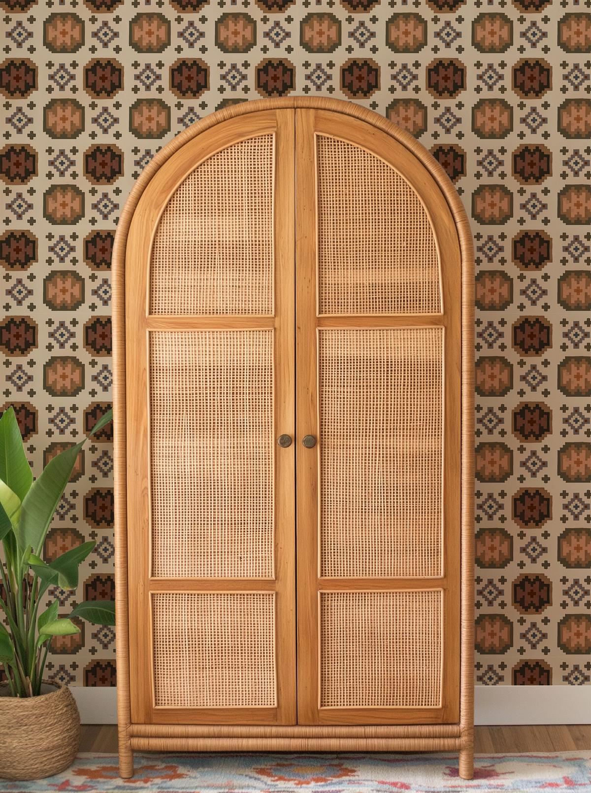 A tall arched wooden cabinet with woven cane panels on the doors, shown in a styled room setting with patterned wallpaper, a potted plant, and a rug.