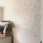 A serene nursery corner featuring wallpaper with a woodland animal theme in beige, showing rabbits, squirrels, and hedgehogs, complemented by a pale pink pig-shaped pillow and fresh flowers.