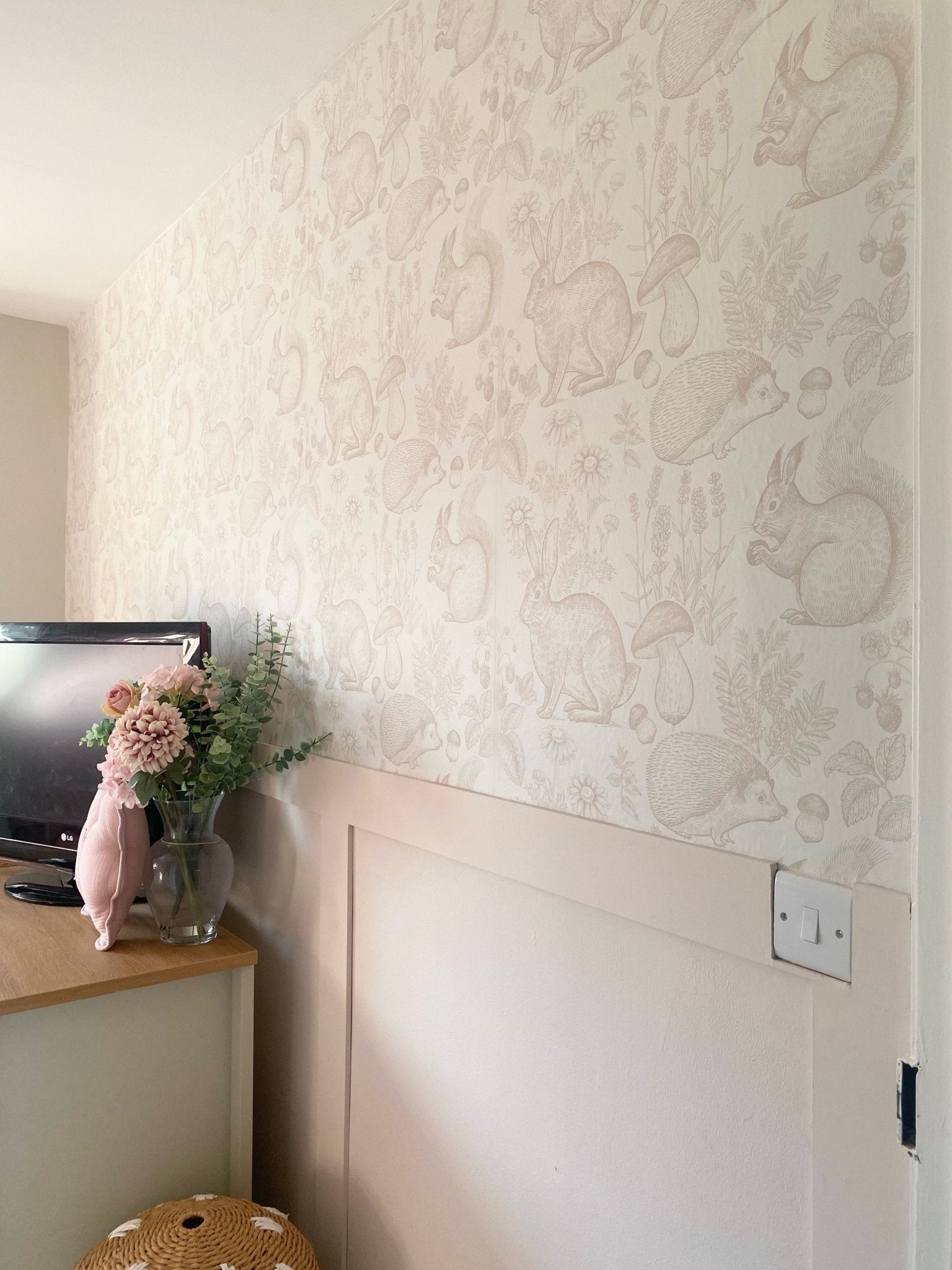 A serene nursery corner featuring wallpaper with a woodland animal theme in beige, showing rabbits, squirrels, and hedgehogs, complemented by a pale pink pig-shaped pillow and fresh flowers.