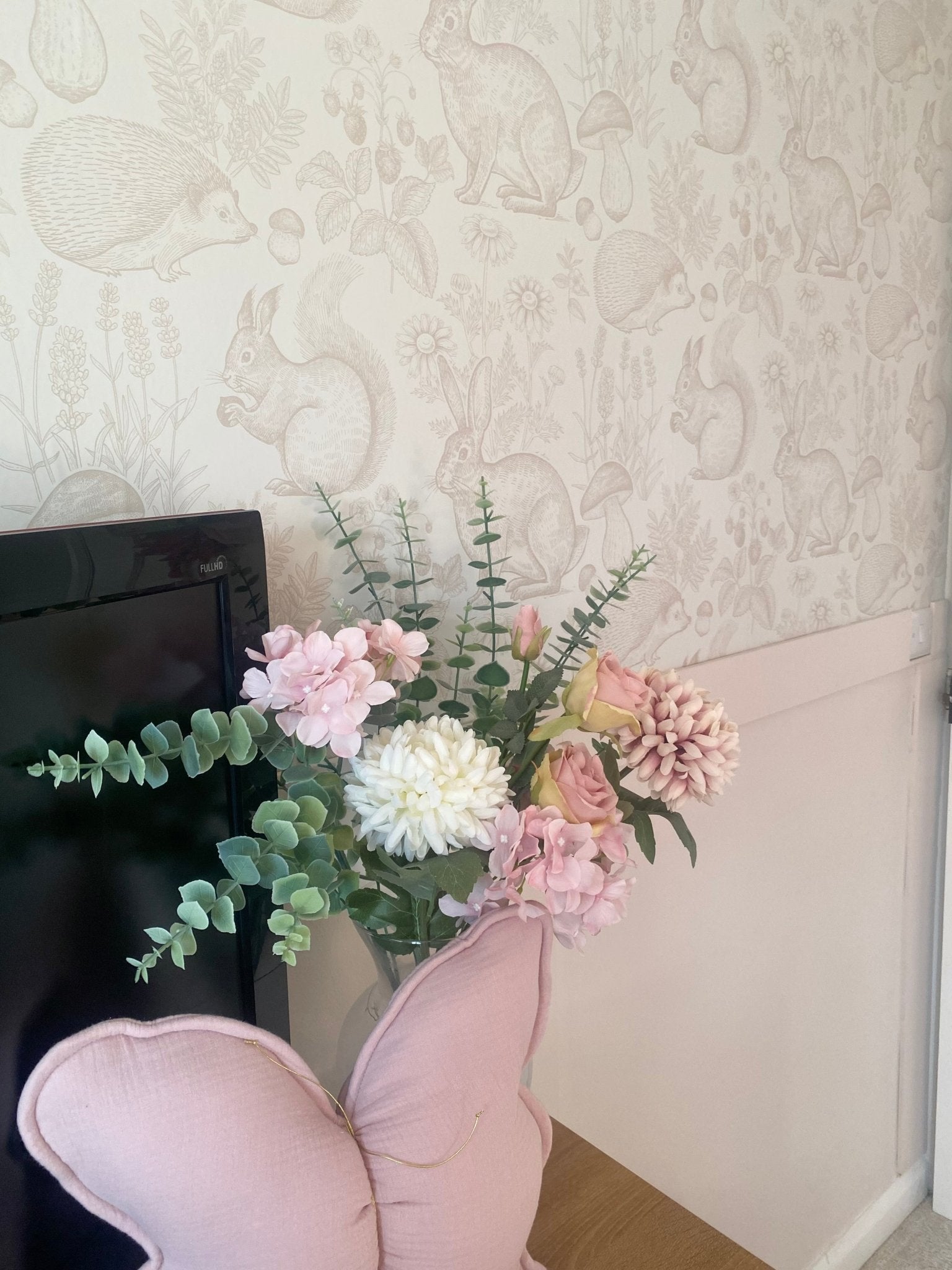 Cozy nursery corner with woodland animal-themed wallpaper in soft beige, adorned with a decorative bouquet and a pink pig-shaped pillow on a small cabinet