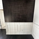 A close-up view of a wall covered in Black Grasscloth wallpaper with white wainscoting below, highlighting the elegant texture and depth of the wallpaper in a room setting.