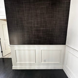 A close-up view of a wall covered in Black Grasscloth wallpaper with white wainscoting below, highlighting the elegant texture and depth of the wallpaper in a room setting.