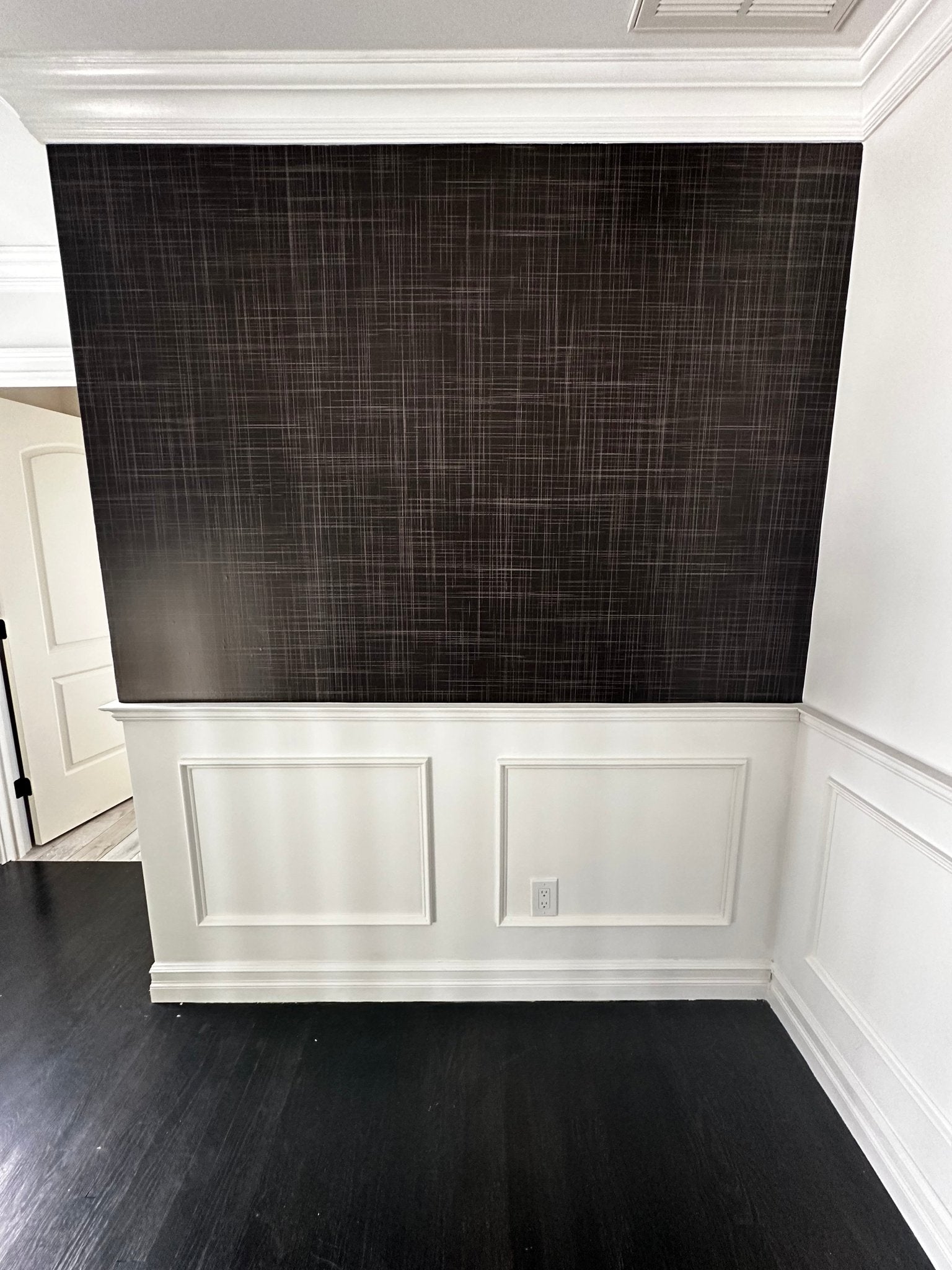 A close-up view of a wall covered in Black Grasscloth wallpaper with white wainscoting below, highlighting the elegant texture and depth of the wallpaper in a room setting.