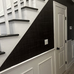 A staircase with white risers and dark steps, featuring Black Grasscloth wallpaper along the stair wall, adding a sleek, textured contrast to the white wainscoting below.