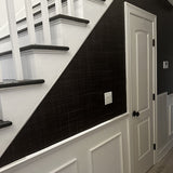 A staircase with white risers and dark steps, featuring Black Grasscloth wallpaper along the stair wall, adding a sleek, textured contrast to the white wainscoting below.