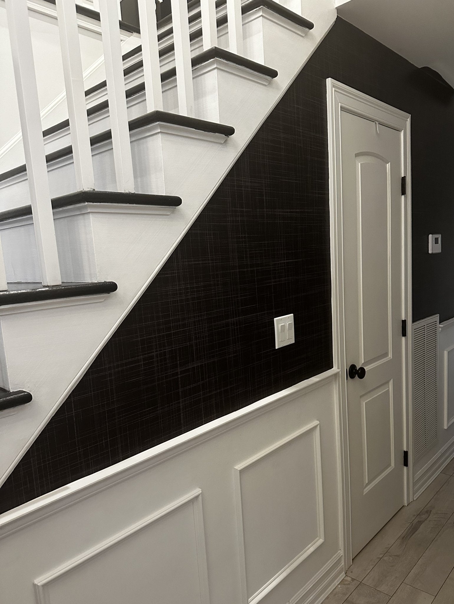 A staircase with white risers and dark steps, featuring Black Grasscloth wallpaper along the stair wall, adding a sleek, textured contrast to the white wainscoting below.