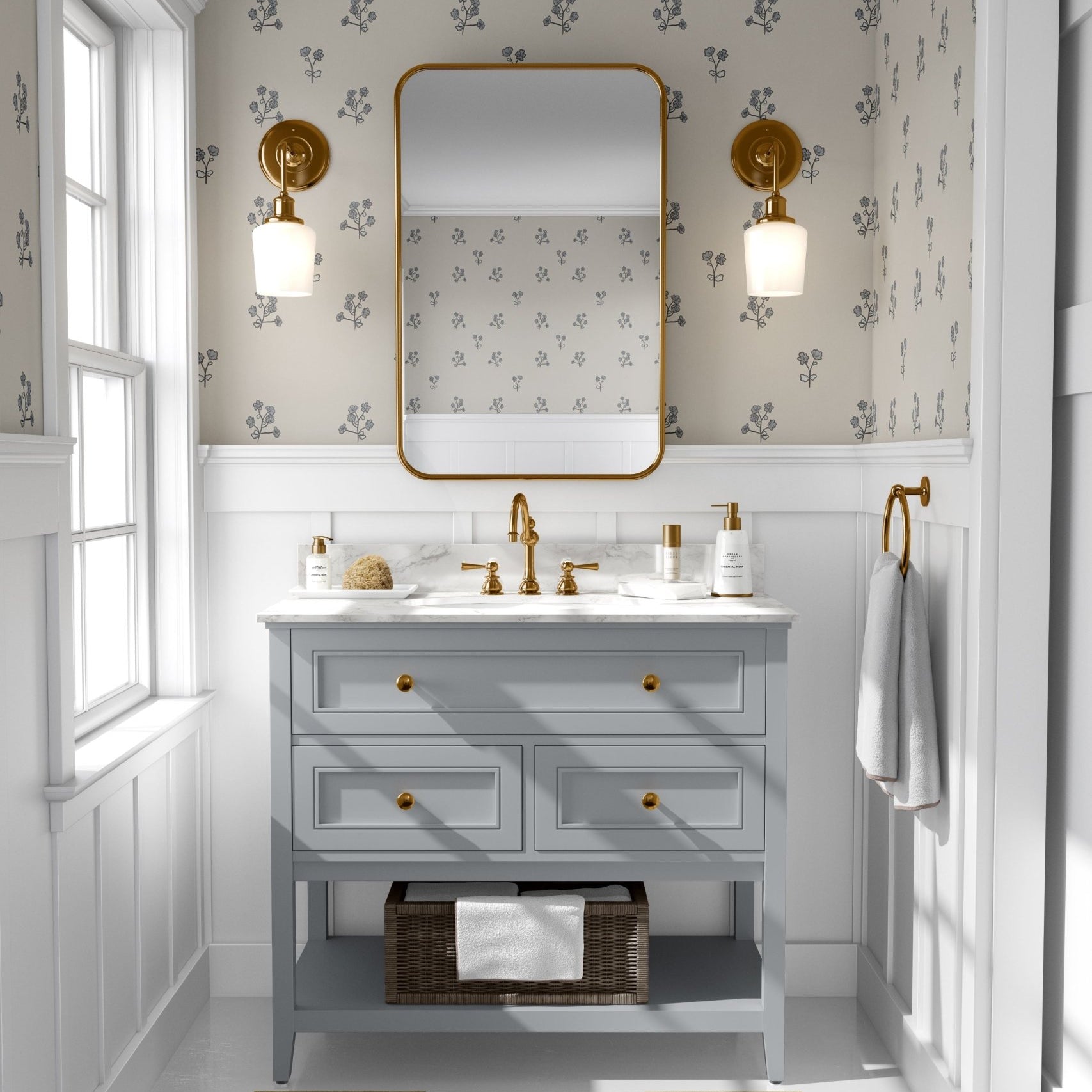 A bright bathroom with walls covered in bluebell flower wallpaper matching the previous image. The room features a light blue vanity with golden knobs, a marble top, golden faucets, and a large mirror with a golden frame.