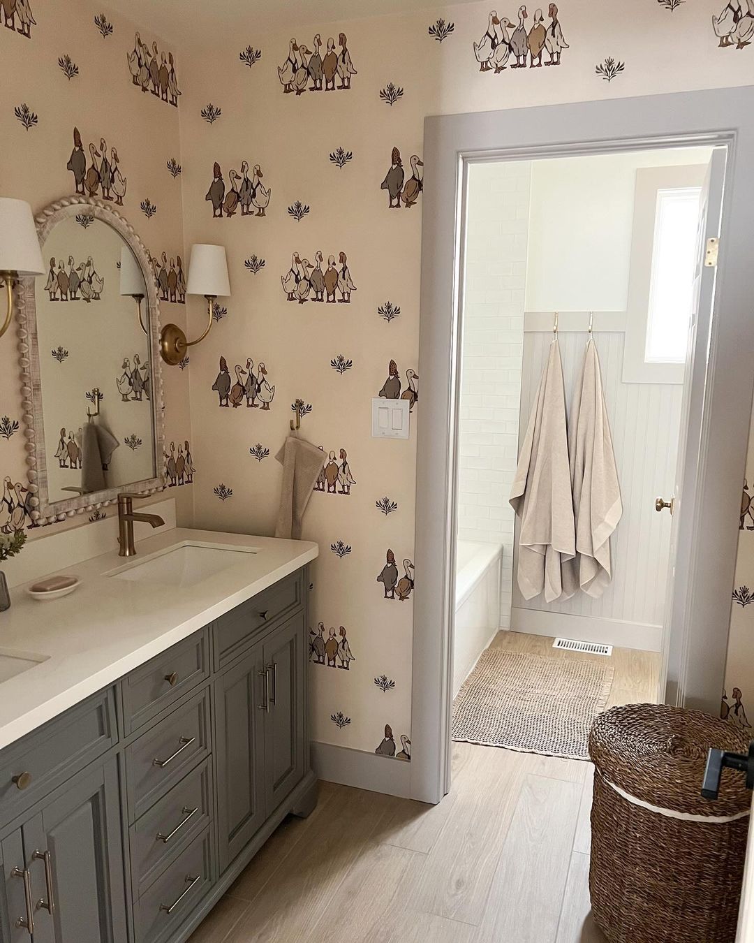 Charming bathroom featuring duck-themed wallpaper with neutral tones. The bathroom includes a classic vanity with a white countertop and gold fixtures, a large mirror with decorative trim, and a door to a bath area with white tile and beige towels