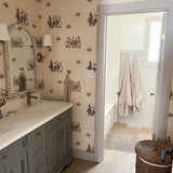 Charming bathroom featuring duck-themed wallpaper with neutral tones. The bathroom includes a classic vanity with a white countertop and gold fixtures, a large mirror with decorative trim, and a door to a bath area with white tile and beige towels