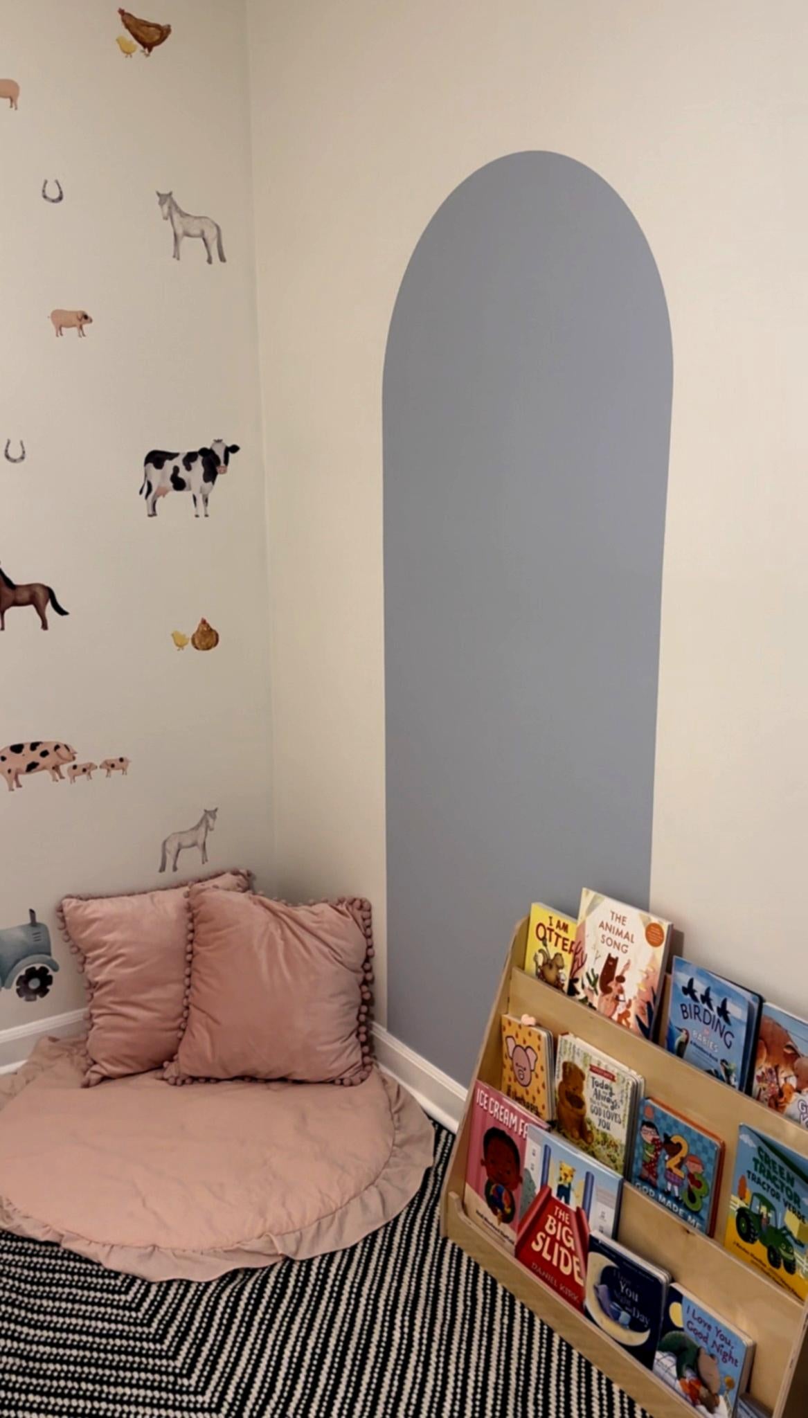 Children's reading nook with a cozy floor pillow and a blue sticker arch on the wall. The wall features playful farm animal decals, and a wooden bookshelf displays various children's books