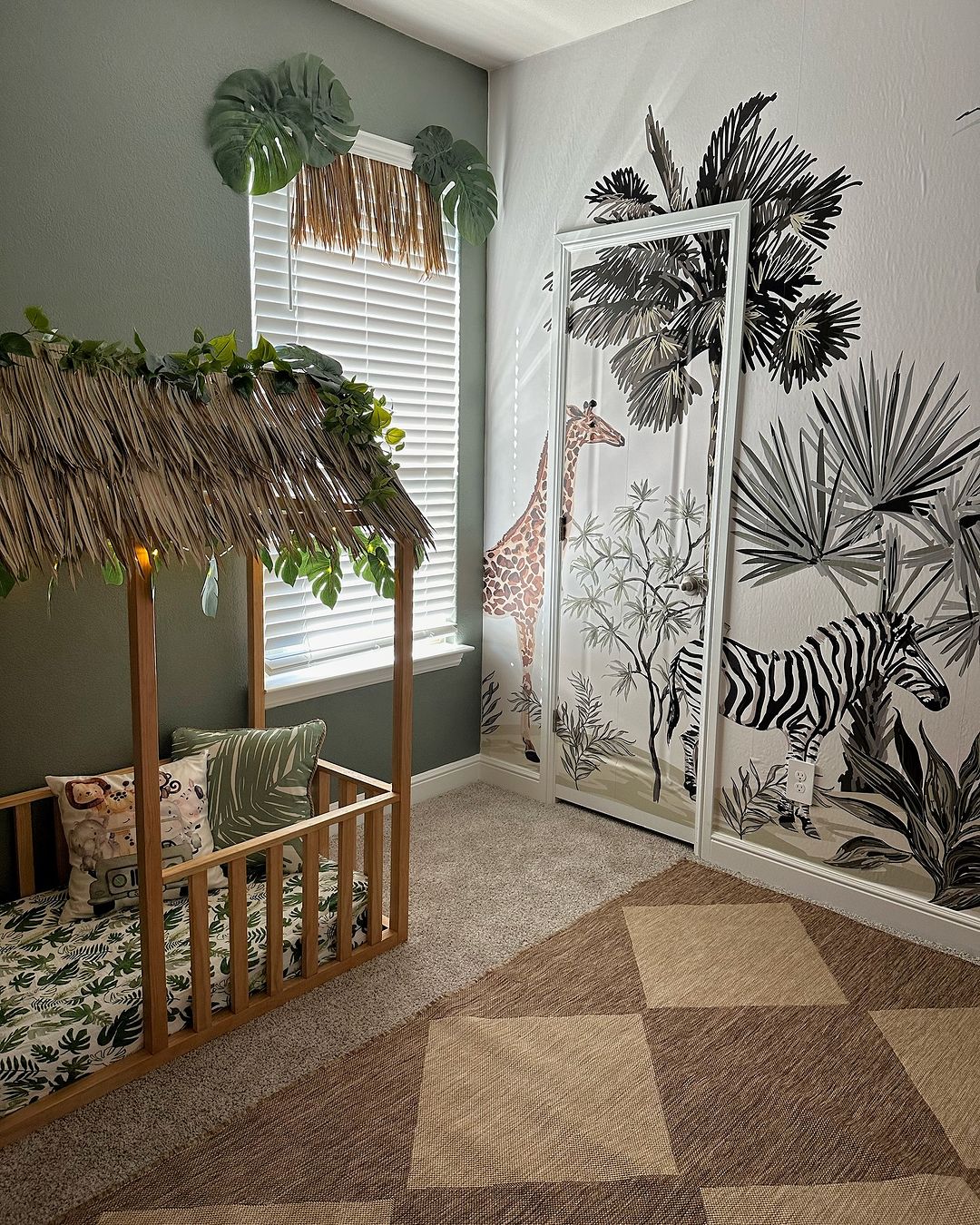 Jungle safari playroom decor with a safari mural showcasing giraffes, zebras, and tropical plants. Wooden crib under a thatched canopy adorned with greenery, creating a tropical, adventurous feel.