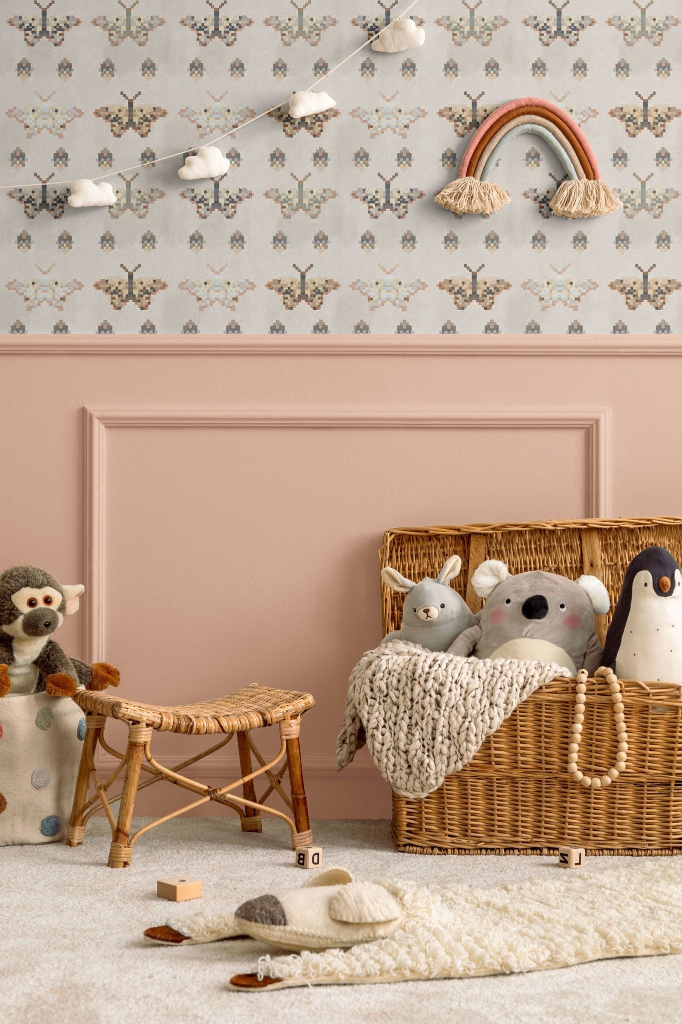 Cozy children's room with mosaic butterfly wallpaper in pastel and earthy tones. The room features a wicker toy basket with plush animals, a rattan stool, and a pink wall with a rainbow decoration and cloud garland, creating a warm, playful space