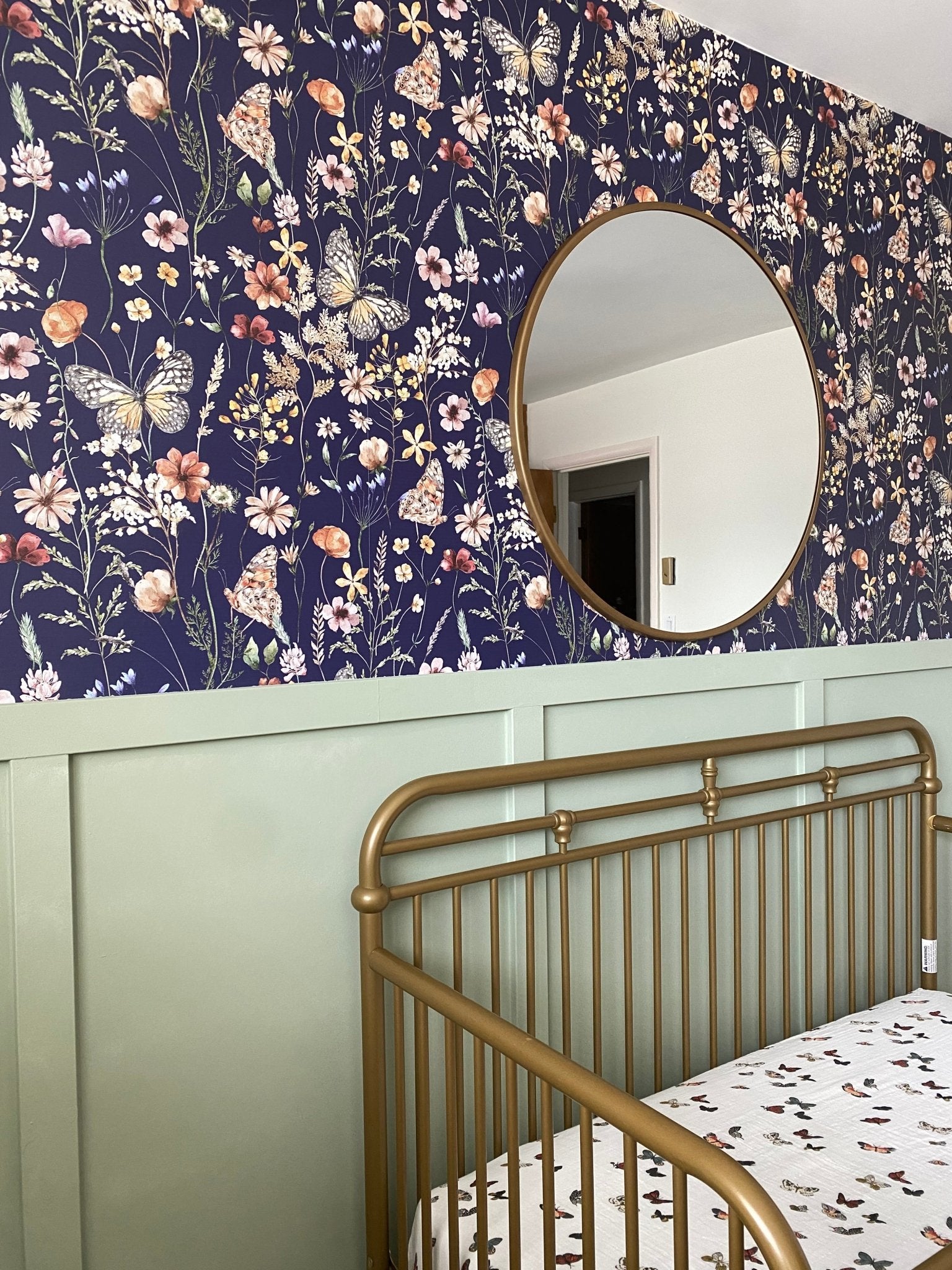  A charming nursery featuring a navy blue wallpaper adorned with detailed floral and butterfly patterns, complemented by a circular mirror and a green half-wall.