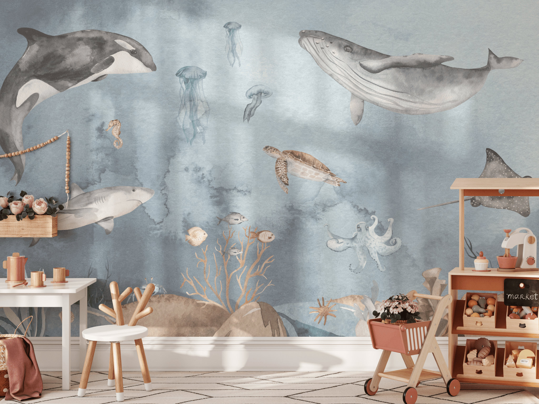A children's playroom featuring an ocean-themed wall mural. The mural showcases a variety of sea creatures including orcas, sharks, whales, jellyfish, sea turtles, and an octopus, all painted in a soft watercolor style. The room is furnished with a small white table, wooden chairs, and a toy cart filled with plush toys and children's books, enhancing the marine nursery decor.