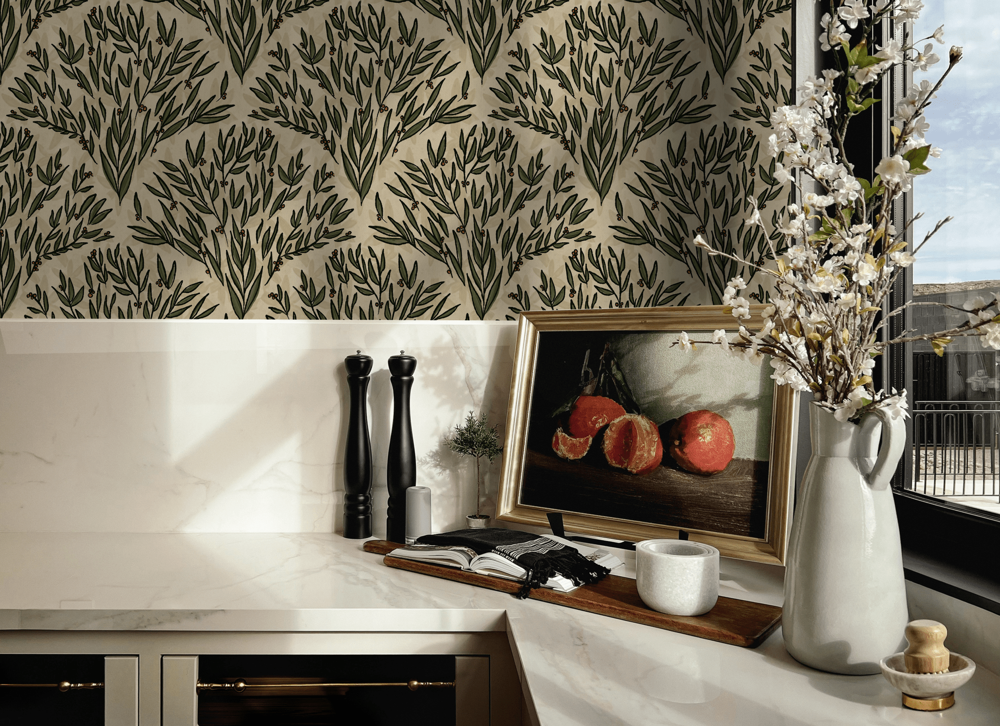 A stylish kitchen corner showcasing olive branch wallpaper as a backdrop. The scene includes marble countertops, a vase with blooming branches, salt and pepper mills, and an artwork of pomegranates.