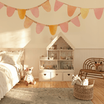 A charming bedroom with a pastel bunting banner wall decal in pink, yellow, and orange hues above a dollhouse. The room features a wooden chair, a soft rug, and a basket of toys, creating a cozy and whimsical setting for kids.