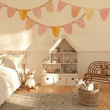 A charming bedroom with a pastel bunting banner wall decal in pink, yellow, and orange hues above a dollhouse. The room features a wooden chair, a soft rug, and a basket of toys, creating a cozy and whimsical setting for kids.