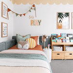 A children's bedroom showcasing a bed with colorful bedding and cushions, a wall-mounted bookshelf filled with books, and framed artwork. A scalloped wallpaper border in a soft peach hue accents the top of the walls, creating a playful atmosphere