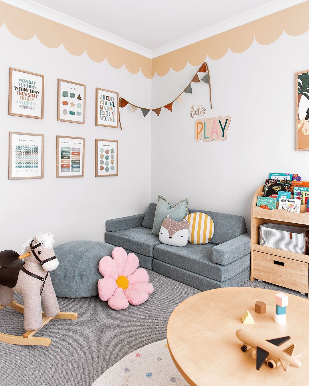 A charming children's playroom with a soft grey sectional sofa, decorative pillows, a wooden rocking horse, and a flower-shaped floor cushion. The room features a playful scalloped wallpaper border in a soft peach color near the ceiling, adding a whimsical touch.