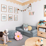 A charming children's playroom with a soft grey sectional sofa, decorative pillows, a wooden rocking horse, and a flower-shaped floor cushion. The room features a playful scalloped wallpaper border in a soft peach color near the ceiling, adding a whimsical touch.